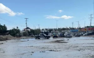 Urban street heavily flooded with muddy water; vehicles navigate through the water and a police pickup is in the foreground.