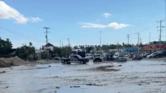 Urban street heavily flooded with muddy water; vehicles navigate through the water and a police pickup is in the foreground.