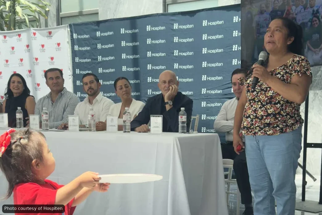 Woman in a floral top speaks into a microphone at a panel discussion, with six panelists seated behind a white table and Hospiten banners in the background.