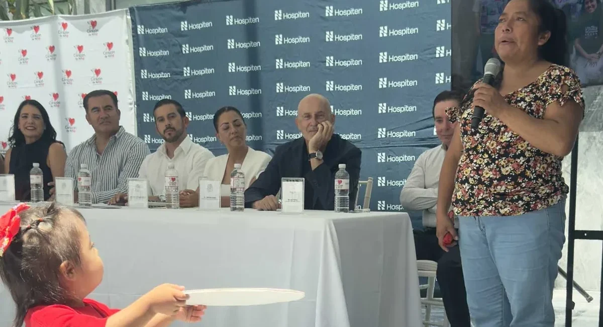 Woman in a floral top speaks into a microphone at a panel discussion, with six panelists seated behind a white table and Hospiten banners in the background.