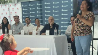 Woman in a floral top speaks into a microphone at a panel discussion, with six panelists seated behind a white table and Hospiten banners in the background.