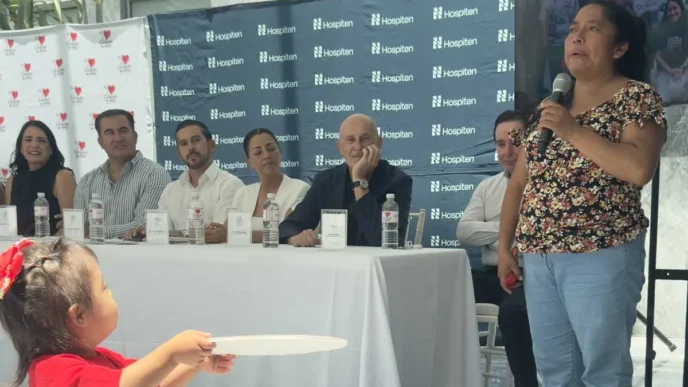 Woman in a floral top speaks into a microphone at a panel discussion, with six panelists seated behind a white table and Hospiten banners in the background.