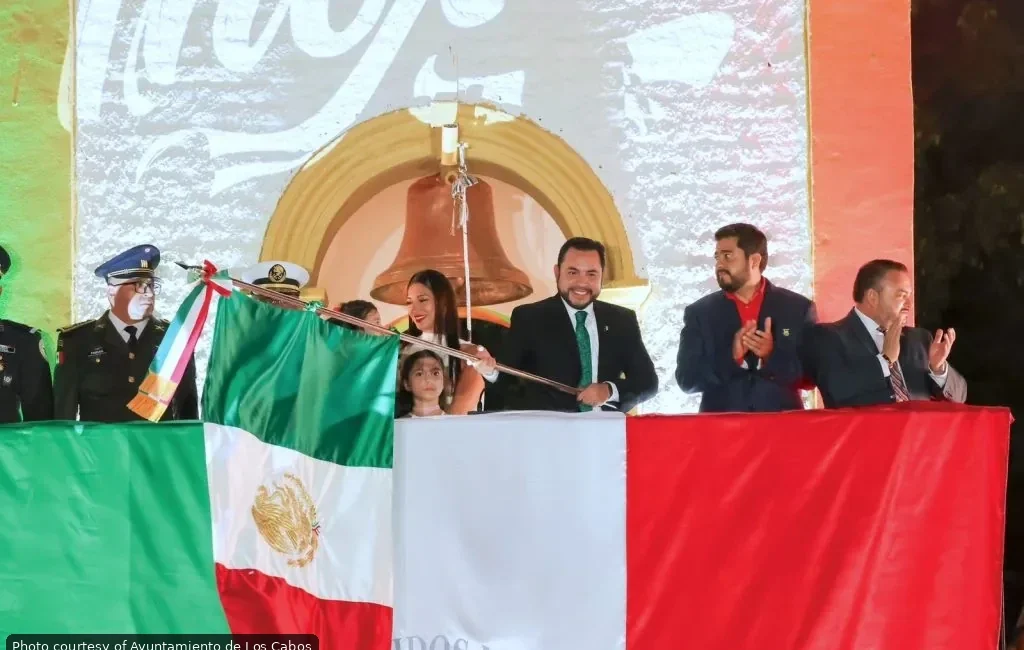 Officials on a stage unveil the Mexican flag during a formal ceremony, with a large backdrop and crowd in the background.