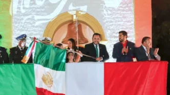 Officials on a stage unveil the Mexican flag during a formal ceremony, with a large backdrop and crowd in the background.