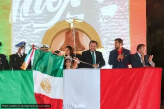 Officials on a stage unveil the Mexican flag during a formal ceremony, with a large backdrop and crowd in the background.