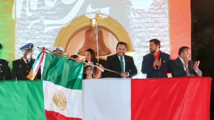 Officials on a stage unveil the Mexican flag during a formal ceremony, with a large backdrop and crowd in the background.