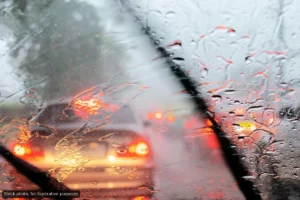 View through a car windshield in heavy rain; wipers sweep as glowing red taillights appear through droplets in traffic ahead.