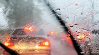 View through a car windshield in heavy rain; wipers sweep as glowing red taillights appear through droplets in traffic ahead.