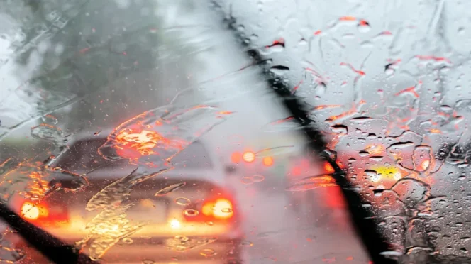 View through a car windshield in heavy rain; wipers sweep as glowing red taillights appear through droplets in traffic ahead.