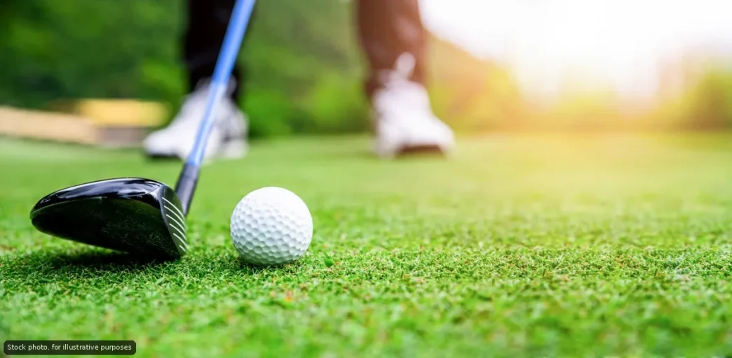 Golf club behind a white golf ball on a green fairway with a golfer blurred in the background.