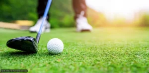 Golf club behind a white golf ball on a green fairway with a golfer blurred in the background.