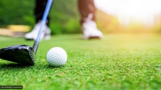 Golf club behind a white golf ball on a green fairway with a golfer blurred in the background.