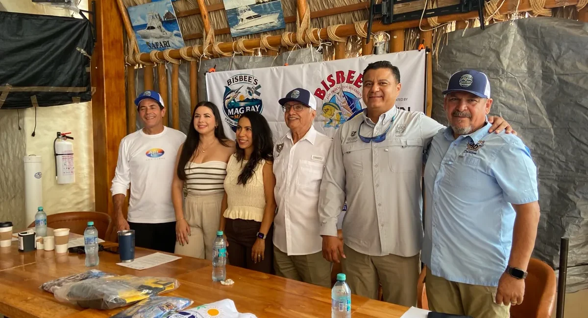 Group of six adults posing for a photo at a Bisbee's event behind a banner, with a wooden table and bottled water in the foreground.