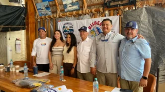 Group of six adults posing for a photo at a Bisbee's event behind a banner, with a wooden table and bottled water in the foreground.