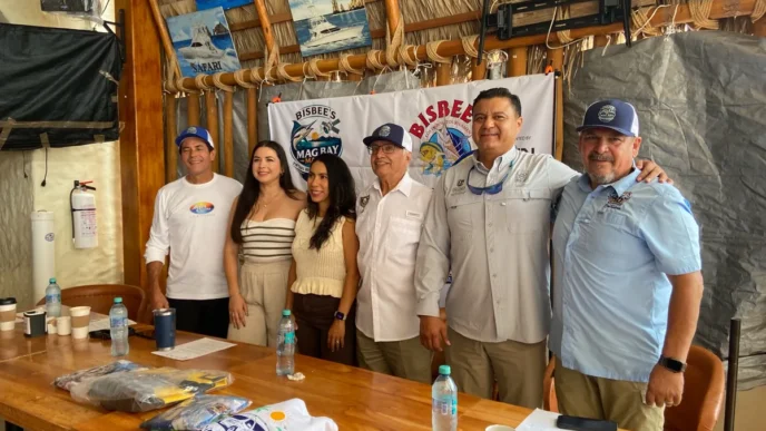 Group of six adults posing for a photo at a Bisbee's event behind a banner, with a wooden table and bottled water in the foreground.