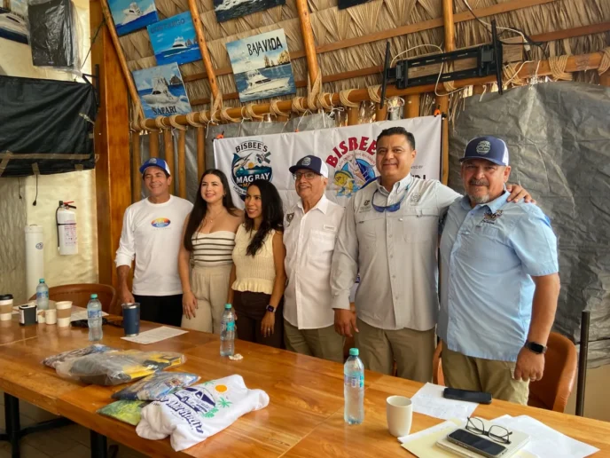 Group of six adults posing for a photo at a Bisbee's event behind a banner, with a wooden table and bottled water in the foreground.