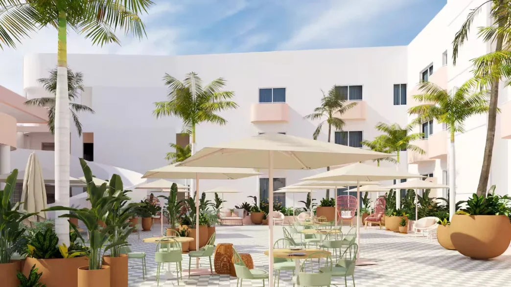 Sunlit hotel courtyard with white buildings, palm trees, and outdoor seating under large umbrellas.