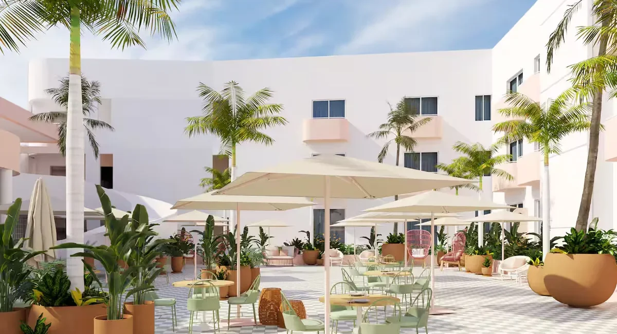 Sunlit hotel courtyard with white buildings, palm trees, and outdoor seating under large umbrellas.