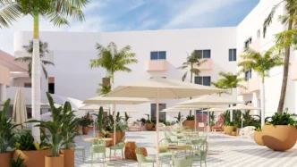 Sunlit hotel courtyard with white buildings, palm trees, and outdoor seating under large umbrellas.