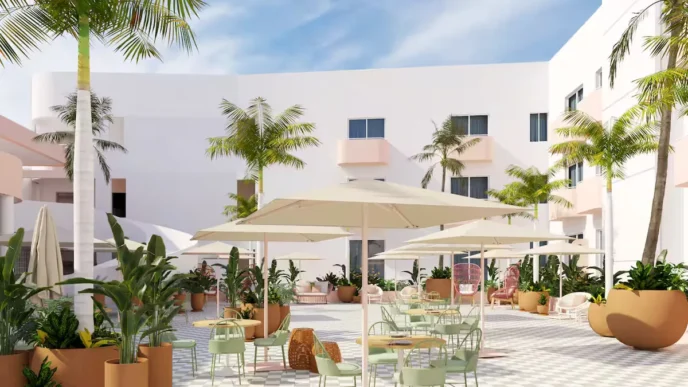 Sunlit hotel courtyard with white buildings, palm trees, and outdoor seating under large umbrellas.
