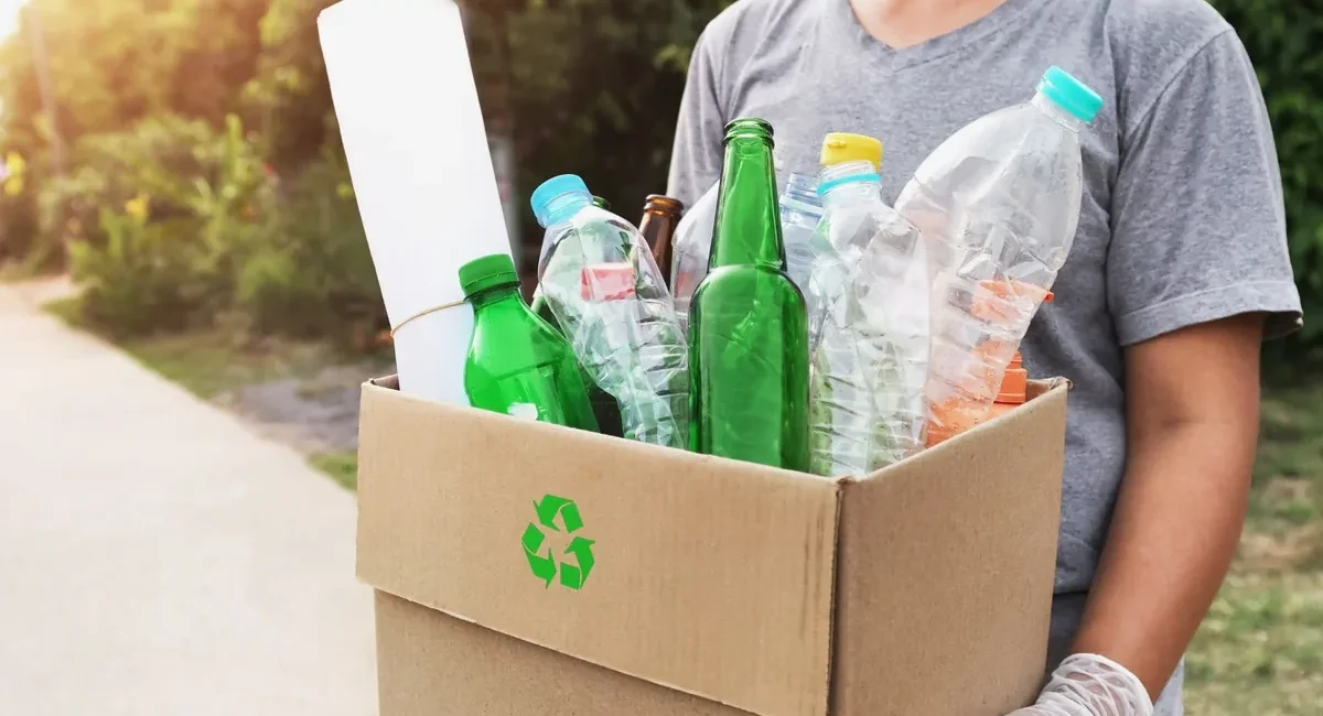 Person in a gray shirt holds a cardboard box full of recyclable plastic bottles for recycling.