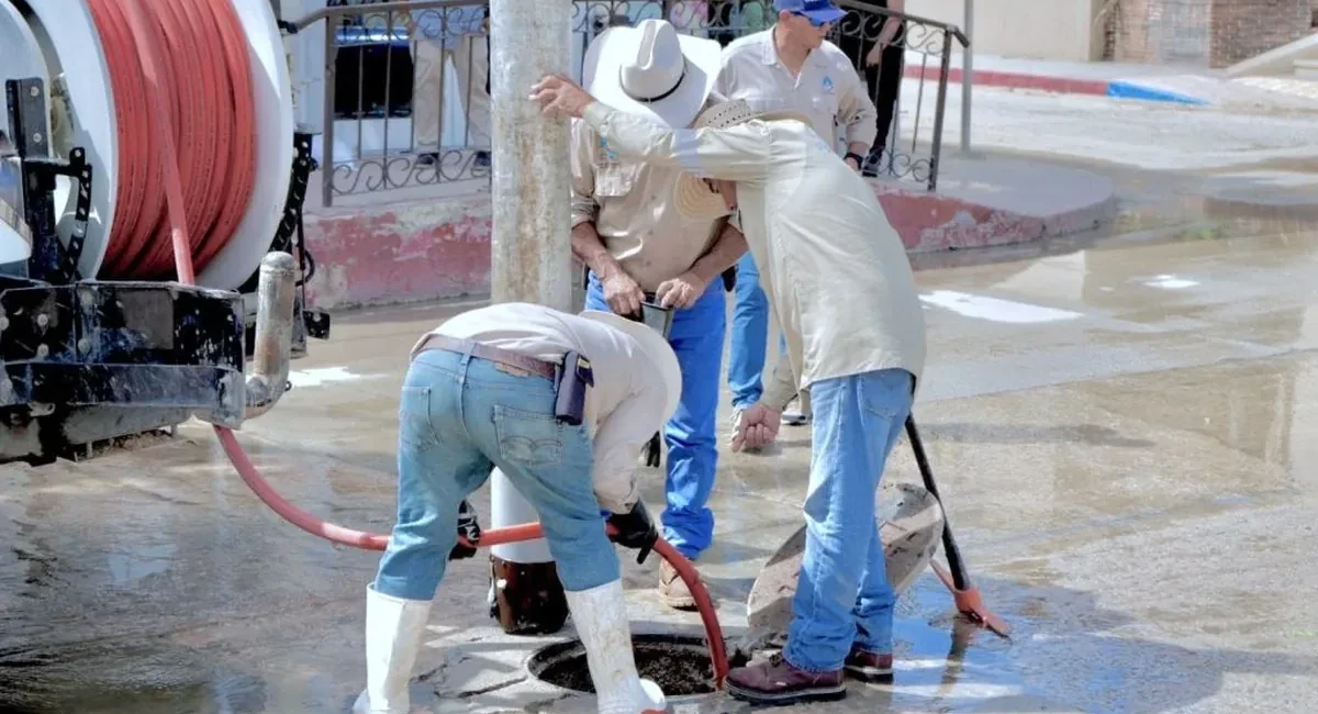 Several workers in wide-brimmed hats and boots repair a street manhole while water pools around them, with a large red hose reel nearby.