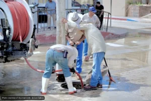 Several workers in wide-brimmed hats and boots repair a street manhole while water pools around them, with a large red hose reel nearby.