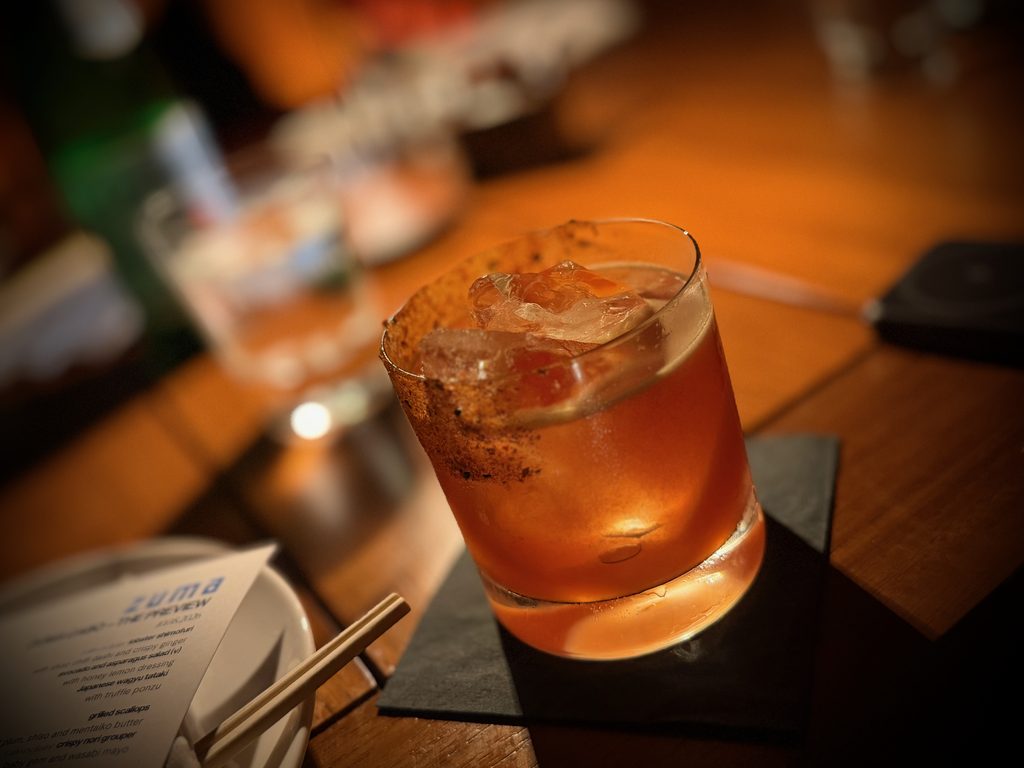 IMG_5537 - Gringo Gazette Close-up of an orange cocktail with ice in a glass on a wooden bar, background blurred.