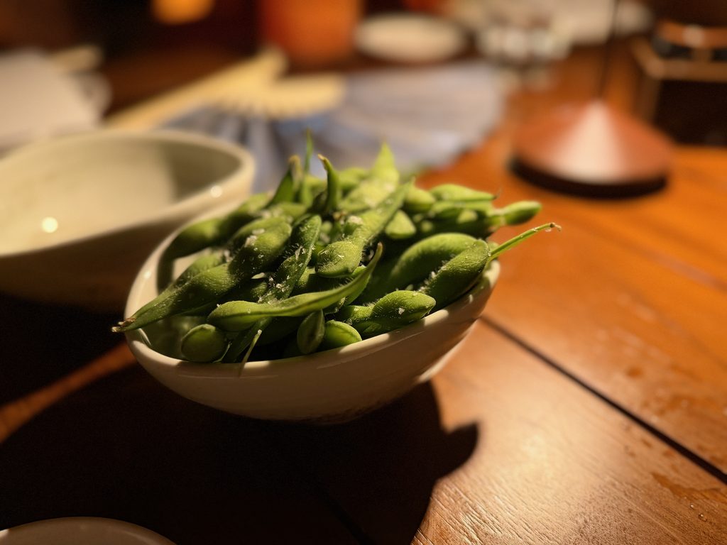 IMG_5543 - Gringo Gazette Close-up of a white bowl filled with fresh green beans on a wooden table, with a shallow depth of field and blurred background.
