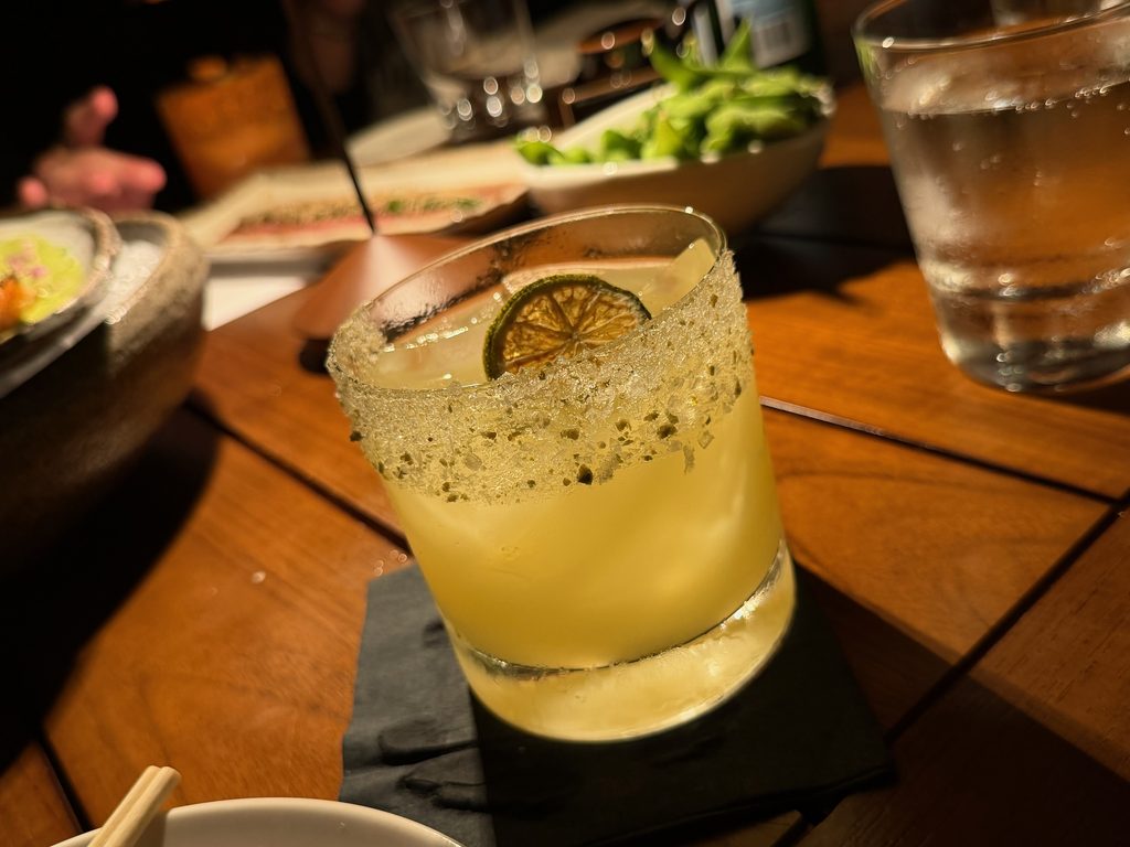 IMG_5555 - Gringo Gazette Salt-rimmed cocktail with lime wheel on a wooden table, blurred restaurant background.