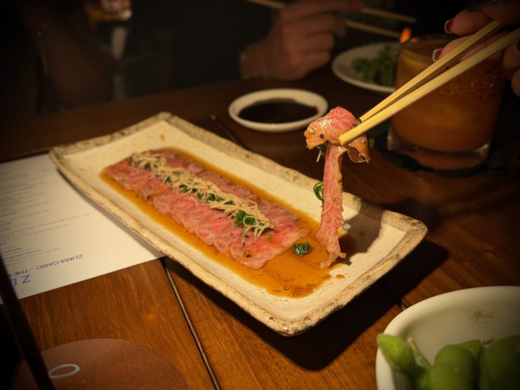 IMG_5559 - Gringo Gazette Thinly sliced beef with orange sauce on a rectangular plate, garnished with greens; chopsticks lift a bite from the dish while diners in dim lighting are in the background.