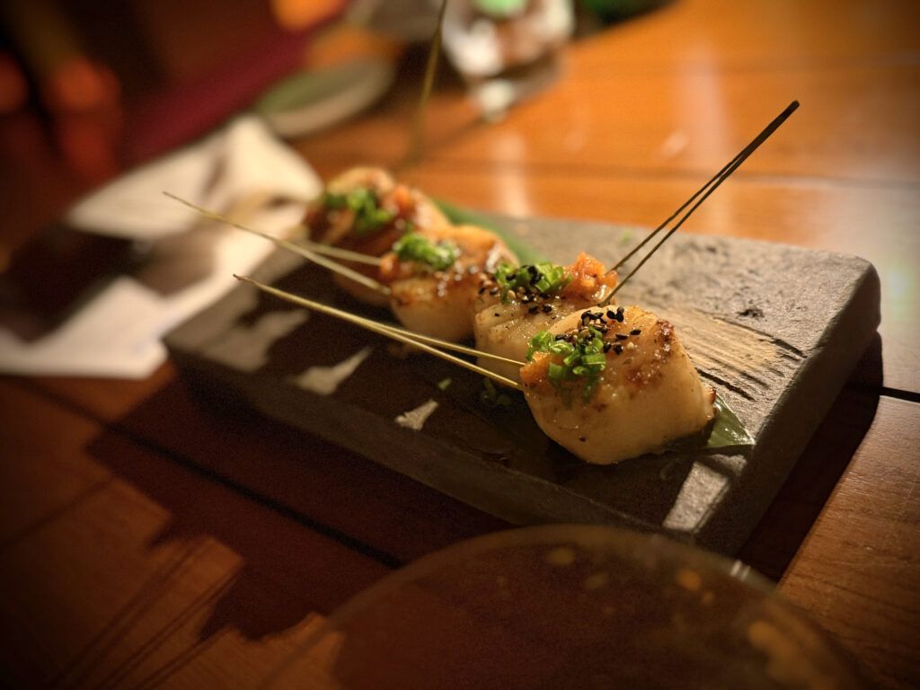 IMG_5575 - Gringo Gazette Skewered grilled scallops topped with chopped herbs and sesame seeds on a dark stone plate