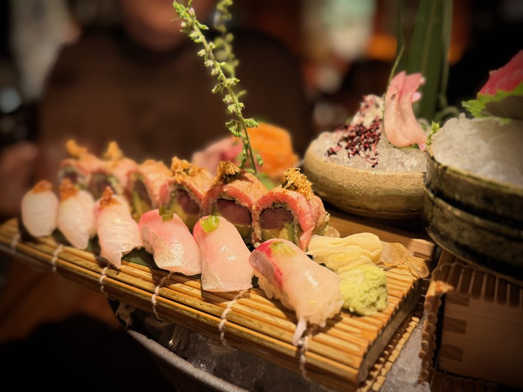 IMG_5587 - Gringo Gazette Close-up of assorted nigiri sushi on a bamboo mat, with fish slices and garnishes like wasabi and herbs.
