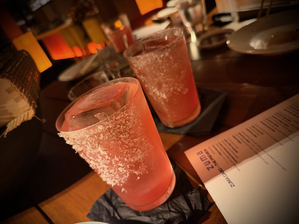 IMG_5591 - Gringo Gazette Two pink cocktails with salted rims on a bar counter, close-up in warm lighting.