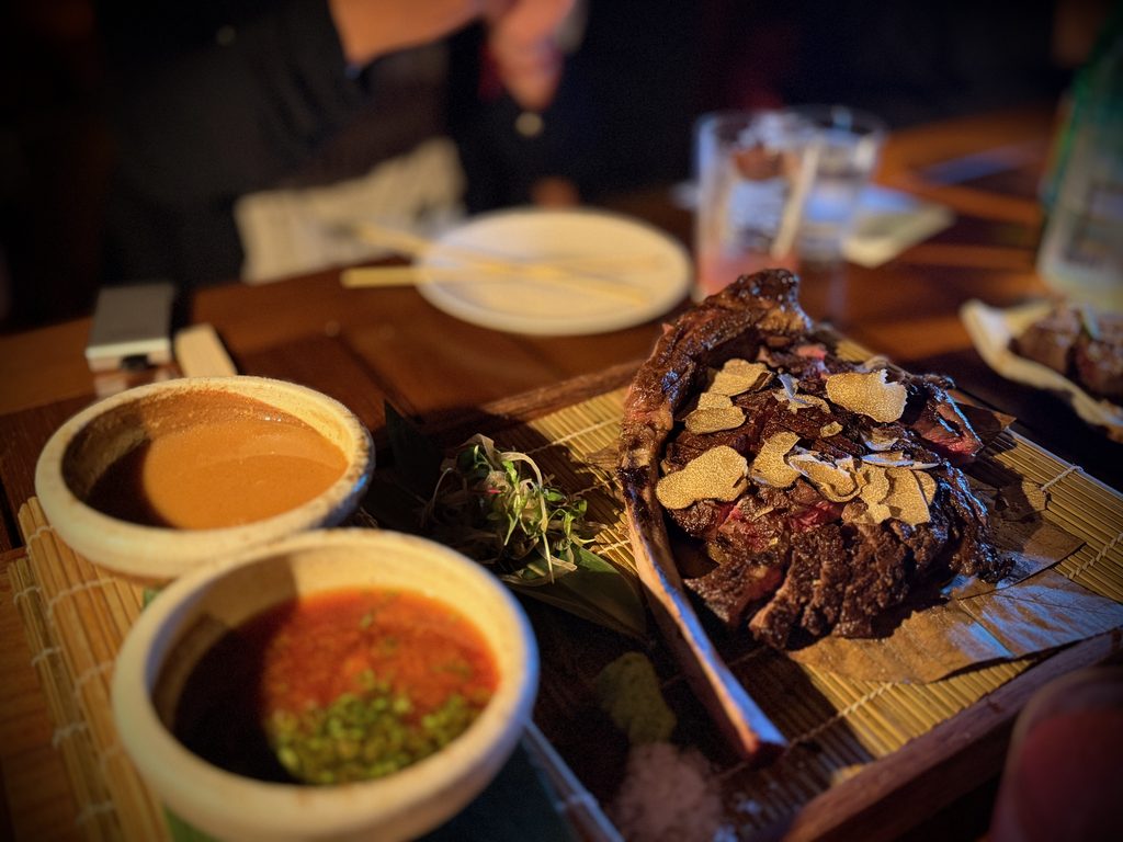 IMG_5596 - Gringo Gazette Close-up of juicy beef topped with truffle shavings on a wooden board, with dipping sauces in the foreground.
