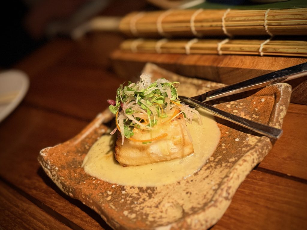 IMG_5600 - Gringo Gazette Seared scallop on toasted bread, topped with shredded vegetables and herbs, surrounded by creamy sauce and a wooden table setting.