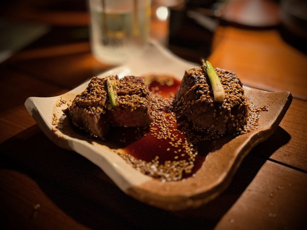 IMG_5602 - Gringo Gazette Sesame-crusted beef cubes on a plate with a glossy sauce and a green onion garnish.