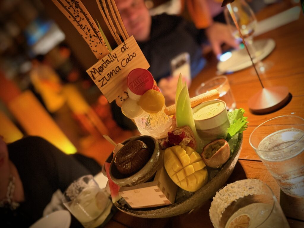 IMG_5609 - Gringo Gazette A close-up of a multi-item appetizer platter with pineapple chunks, citrus slices, and decorative signs, served on a wooden board in a dimly lit setting.