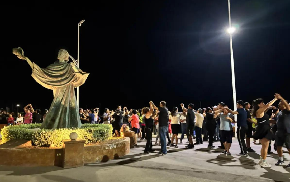 Nighttime scene of a statue in a circular plaza with many people dancing in pairs around it