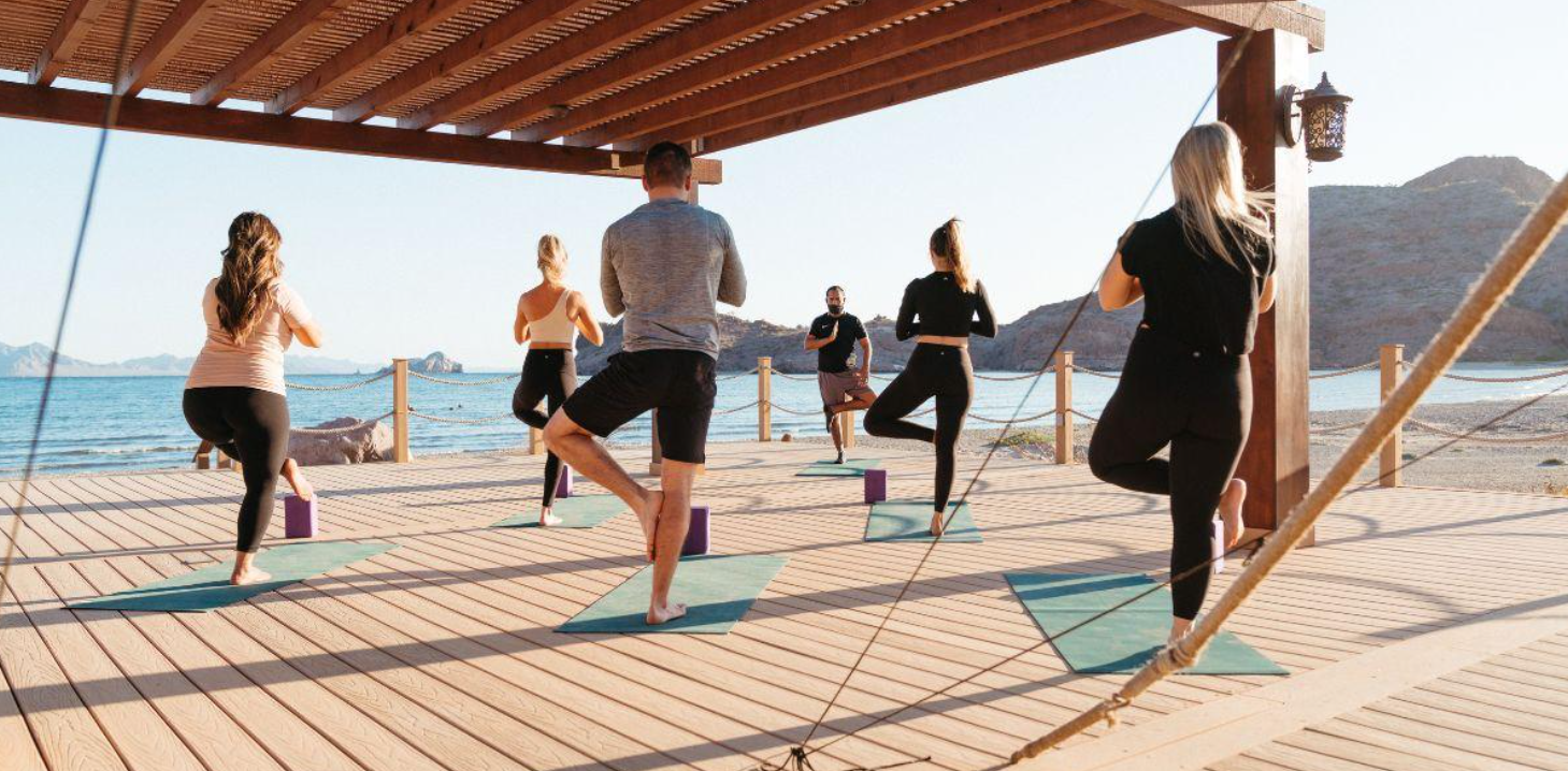 Group of people doing tree pose on yoga mats on a wooden deck by the sea with mountains in the background.
