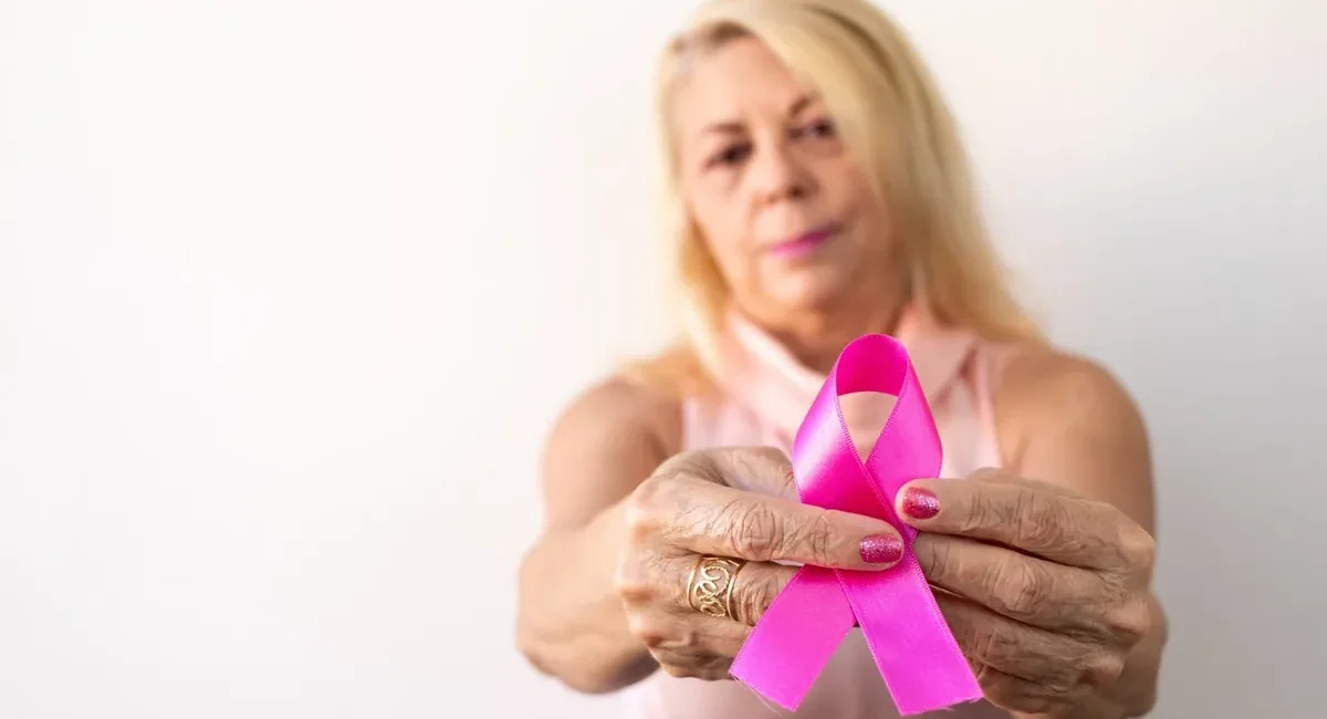 Older woman holding a pink breast cancer awareness ribbon toward the camera, symbolizing support.