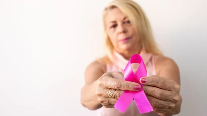 Older woman holding a pink breast cancer awareness ribbon toward the camera, symbolizing support.