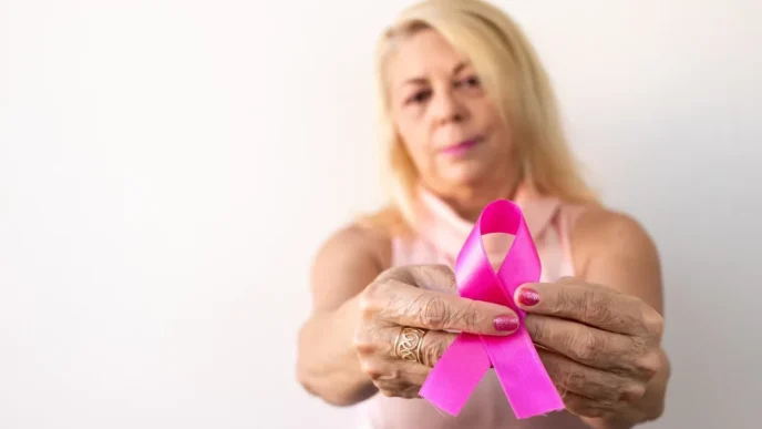 Older woman holding a pink breast cancer awareness ribbon toward the camera, symbolizing support.