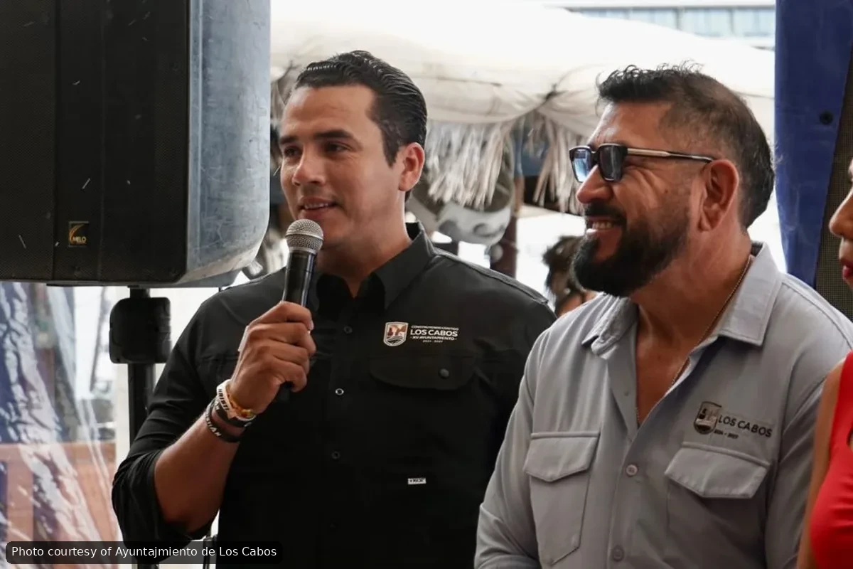 Man in a black Los Cabos shirt speaks into a microphone at an outdoor event, a smiling man in a light gray shirt stands beside him.