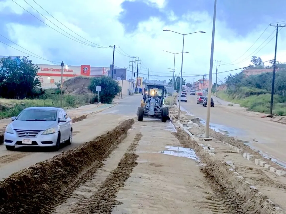Road under construction with a road roller spreading dirt while a silver car passes by on the left.