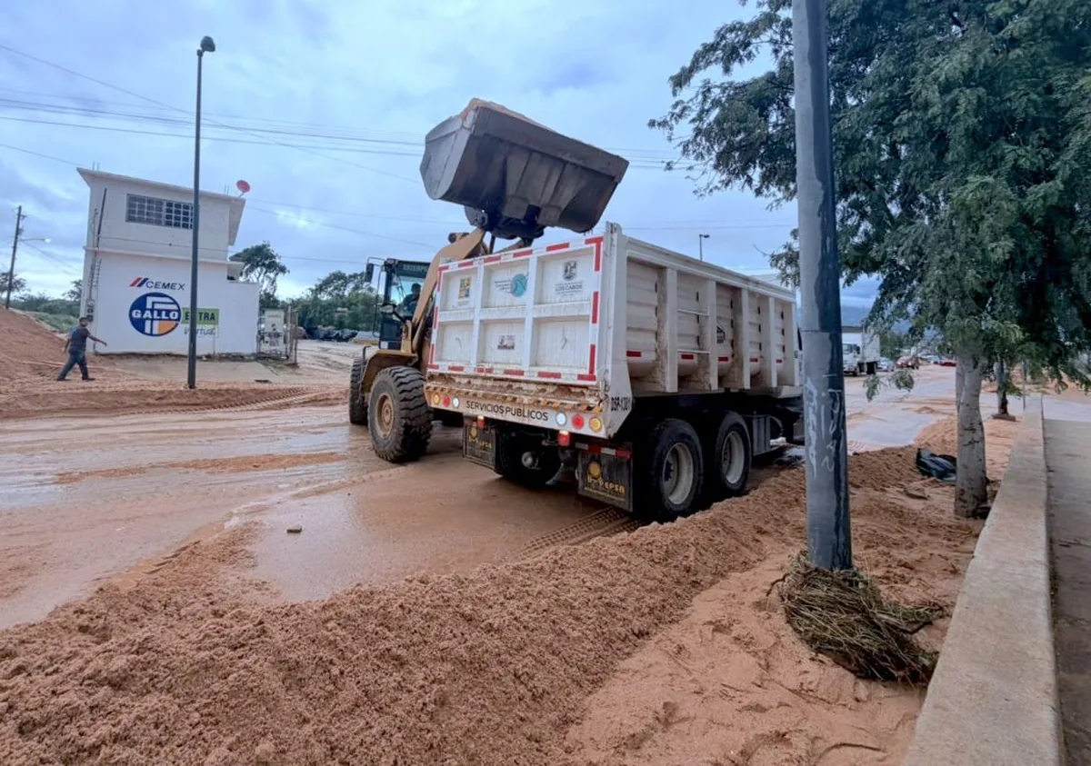 Dump truck unloading sand on a muddy street at a construction site, with a loader’s bucket raised nearby and a Cemex building in the background.