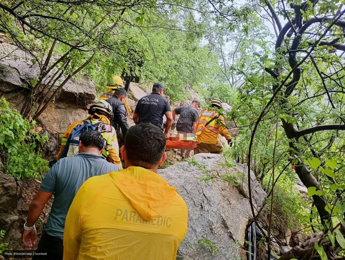 Emergency responders in yellow jackets carry a stretcher up a rocky, forested hillside.