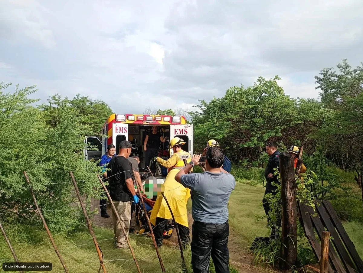 Emergency medical responders load a patient into an EMS ambulance in a rural, fenced field while bystanders observe and a photographer records the scene.