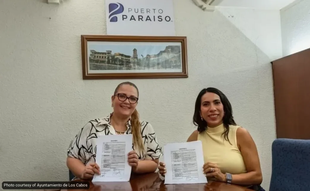 Two women sit at a table holding printed documents, smiling at the camera inside an office with a Puerto Paraiso sign on the wall.