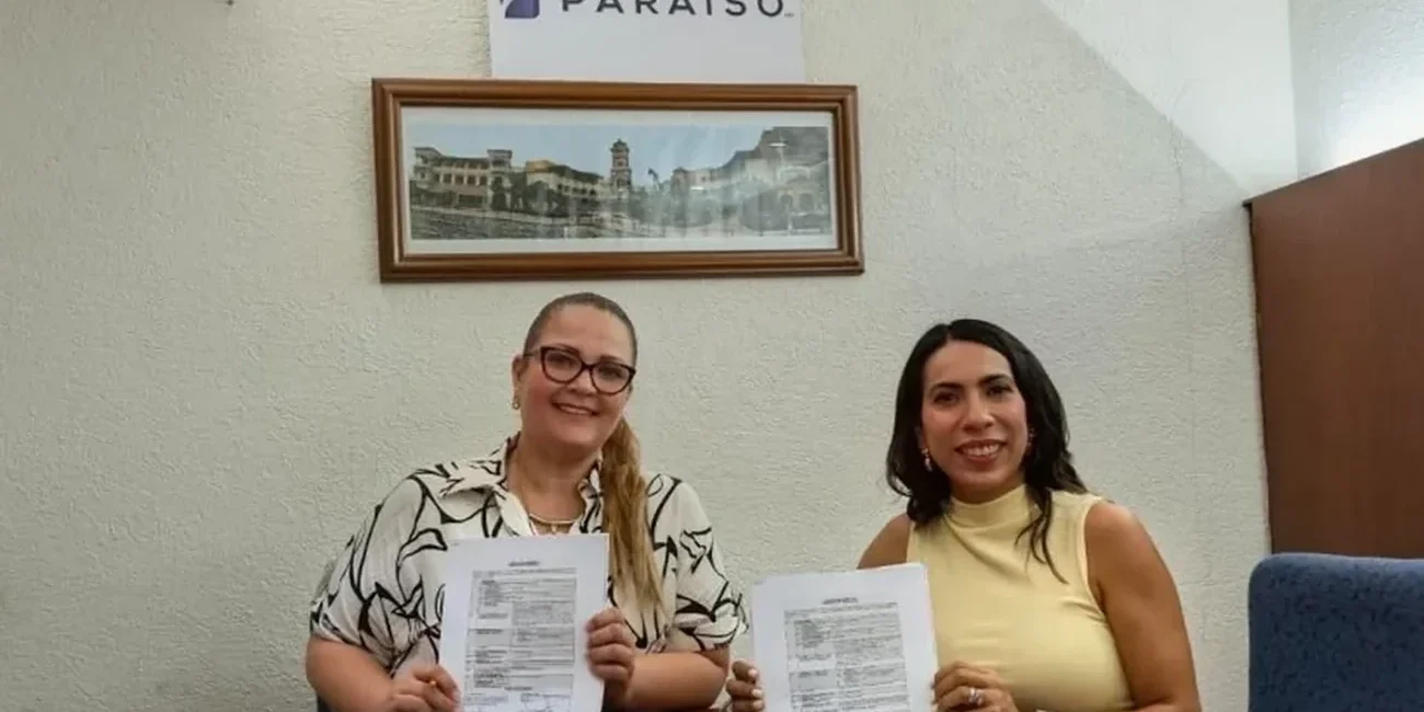 Two women sit at a table holding printed documents, smiling at the camera inside an office with a Puerto Paraiso sign on the wall.